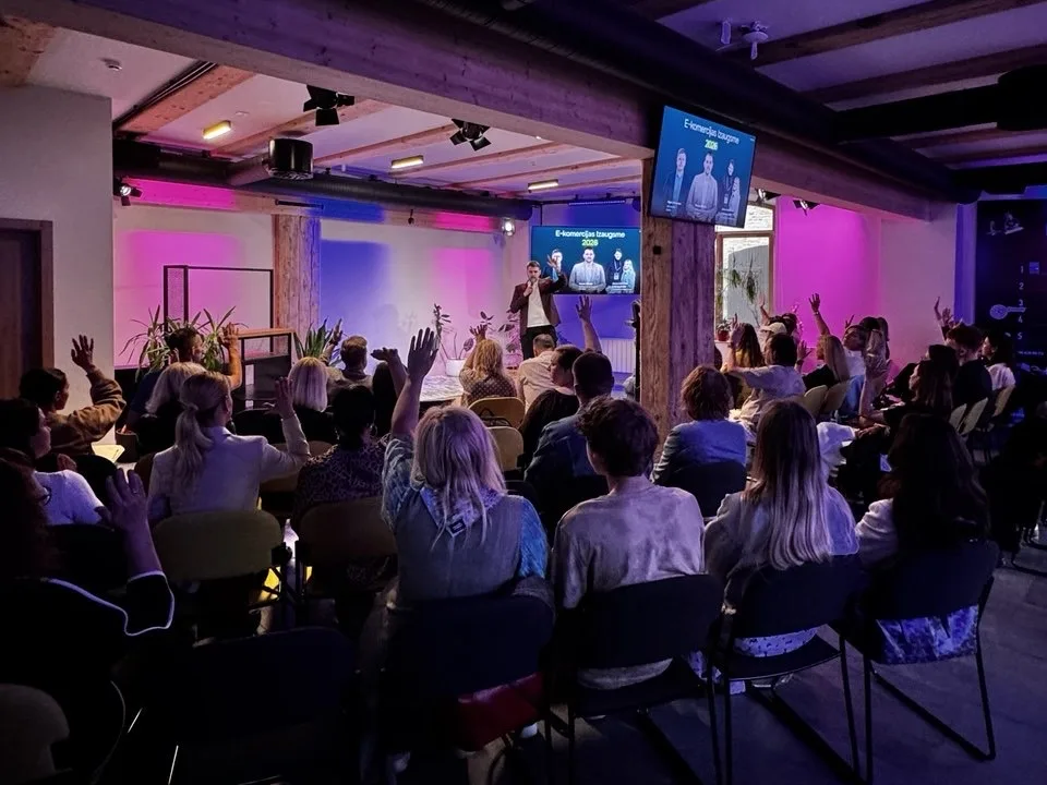 A speaker with a microphone presenting an eCommerce growth talk to a full room, with many people in the audience raising their hands, in a venue lit in purple and pink